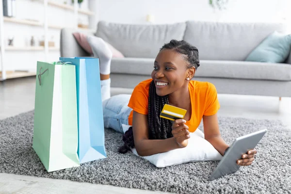 Shopaholism y e-commerce. Señora negra con tarjeta de crédito de compras en línea a través de ordenador portátil, acostado en el suelo con bolsas en el interior —  Fotos de Stock