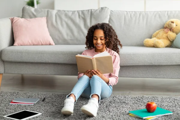Youth and literature. Smiling african american gen z girl holding and reading paper book, sitting on the floor carpet