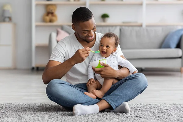 Feliz jovem preto pai colher alimentando seu adorável bebê filho em casa — Fotografia de Stock
