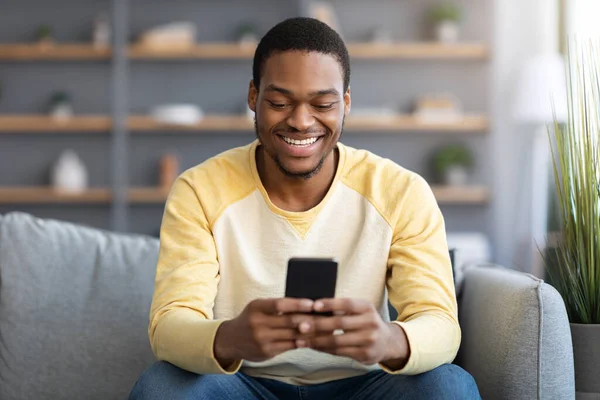 Улыбающийся черный парень сидит на диване, используя смартфон — стоковое фото