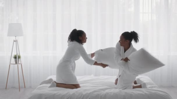 Volwassen en tiener afrikaanse amerikaanse zusters met kussen vechten en lachen op bed thuis, rotzooien en lachen — Stockvideo