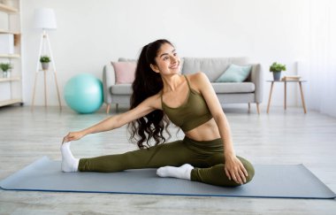 Yurtiçi sporlar. Spor kıyafetli atletik Hintli kadın yoga minderinde spor yapıyor, evde bacaklarını açıyor.