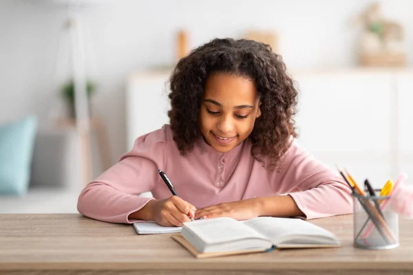 Heimatpädagogisches Konzept. Nettes afrikanisches amerikanisches Schulmädchen macht Hausaufgaben und lächelt, sitzt am Tisch mit Buch — Stockfoto