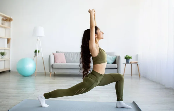 Häusliches Yoga unter Quarantäne. Seitenansicht der sportlichen jungen indischen Dame, die Ausfallschritt macht und zu Hause im Asana-Krieger steht — Stockfoto