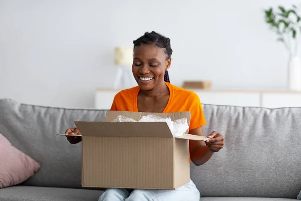 Conceito de compras e entrega online. Alegre mulher negra unboxing pacote de papelão, satisfeito com a compra em casa — Fotografia de Stock