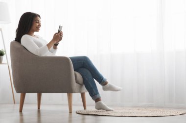 Neşeli Afrikalı Amerikalı kadın evde akıllı telefon kullanıyor, internette sohbet ediyor, koltukta oturuyor, yan manzara, boş alan.