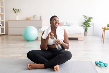 Spor kıyafetli şişman zenci kadın evde bir şişe temiz suyla spor minderinde oturuyor.