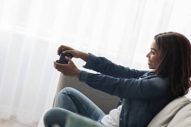 Hafta sonu eğlencesi. Neşeli Afro-Amerikalı kadın çevrimiçi video oyunları oynuyor, elinde joystick, koltukta oturuyor, yandan bakıyor.