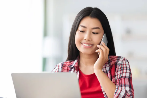 Trabajo a distancia. Feliz joven asiática dama usando el ordenador portátil y hablando por teléfono —  Fotos de Stock