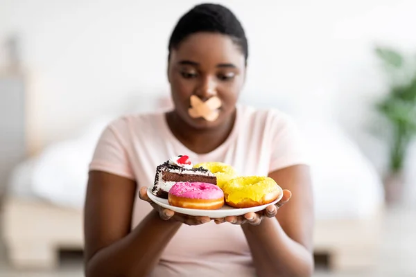 Diet restrictions. Curvy black woman with adhesive bandage on her mouth looking at plate of sweets in hunger at home