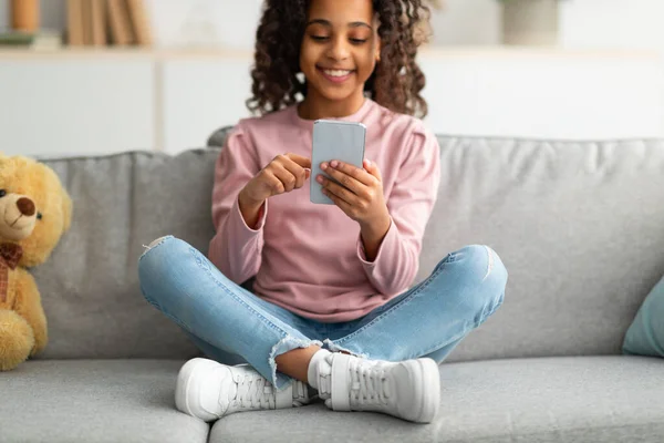 Conceito de jogo móvel. Feliz menina adolescente afro-americana usando smartphone, navegar na internet, sentado no sofá, close-up — Fotografia de Stock