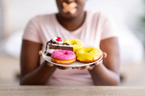 Closeup of curvy black lady holding plate of sweets, having adhesive bandage on her mouth, keeping on weight loss diet