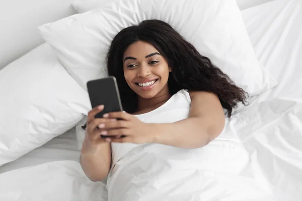 Feliz joven afroamericana dama acostada en la cama y tener videollamada en el teléfono inteligente, acostada en la cama bajo una manta blanca — Foto de Stock