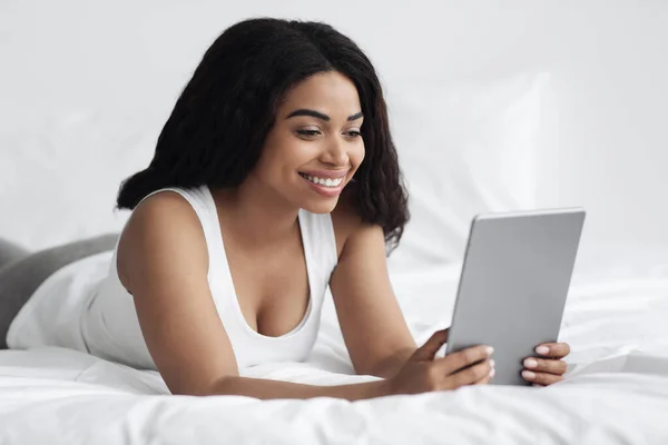 Concepto de tiempo libre y pasatiempo de fin de semana. Feliz dama negra sosteniendo tableta digital, acostado en la cama, espacio libre — Foto de Stock