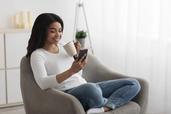 Tiempo libre. Feliz señora afroamericana usando smartphone, sentado en sillón y bebiendo café, espacio libre — Foto de Stock