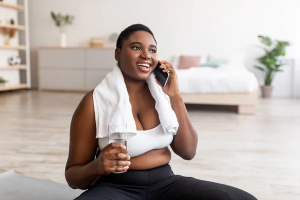 Übergewichtige schwarze Dame mit Wasserglas spricht in der Trainingspause auf dem Smartphone — Stockfoto