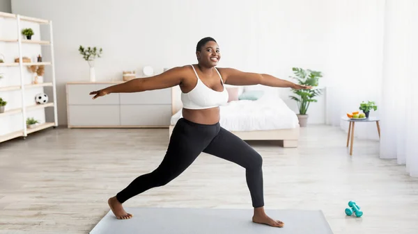 Styrka yoga för viktminskning. Curvy svart dam står i krigare asana, tränar på sportmatta hemma, panorama — Stockfoto