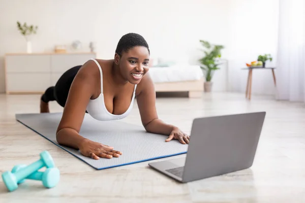 Plump black lady exercising at home, following personal trainers online instructions on laptop, standing in plank pose