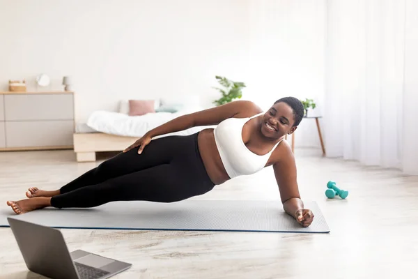 Deportes en línea para bajar de peso. Mujer negra con sobrepeso haciendo ejercicios delante de la computadora portátil, de pie en la tabla lateral en casa —  Fotos de Stock