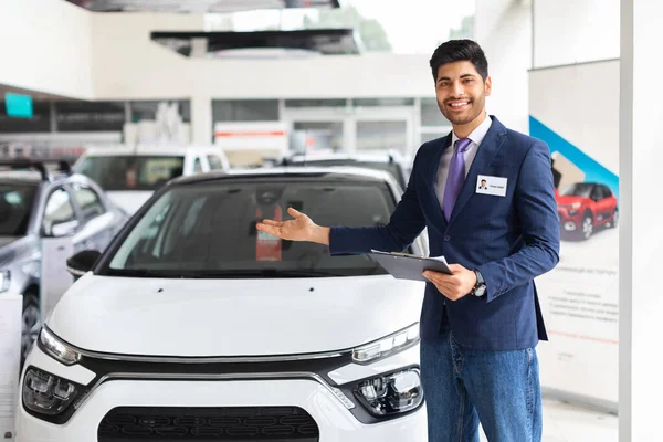Jóképű, fiatal eladó áll a kereskedésben, bemutatja autó — Stock Fotó