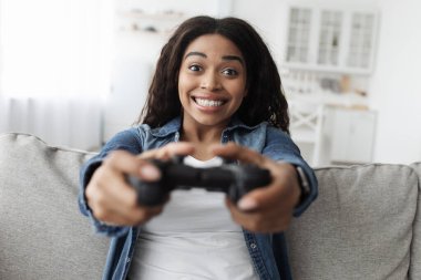 Hafta sonu tatili. Coşkulu Afrikalı Amerikalı kadın joystick kullanıyor, video oyunları oynuyor, evdeki kanepede oturuyor.