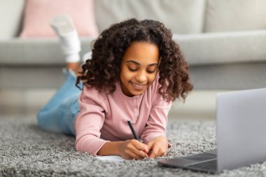 Ev ödevi. Mutlu Afro-Amerikalı kız evde internetten ders çalışıyor dizüstü bilgisayarın önünde notlar alıyor, halının üzerinde yatıyor.
