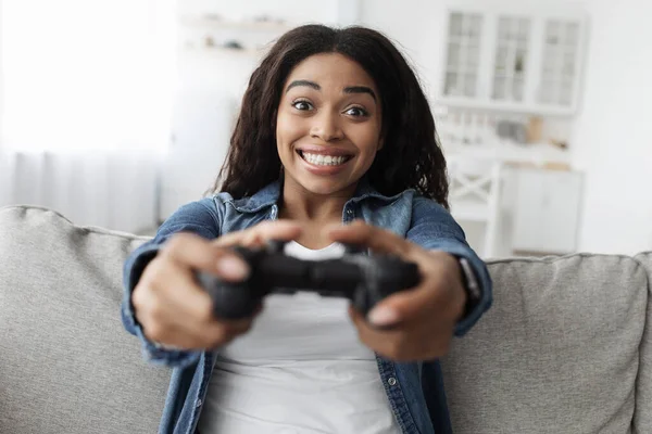 Hétvégi szabadidő. Túlságosan boldog afro-amerikai nő használ joystick és videojátékok, ül a kanapén otthon — Stock Fotó