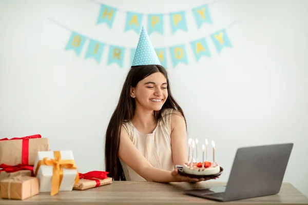 Şenlik şapkalı Hintli genç kız internette doğum günü partisi veriyor, dizüstü bilgisayarında hediye kutuları ve pastayla oturuyor. — Stok fotoğraf