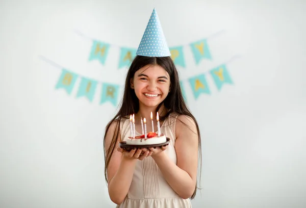 Porträtt av indiska tonåring flicka i festlig hatt håller födelsedagstårta med ljus, ler mot kameran inomhus — Stockfoto