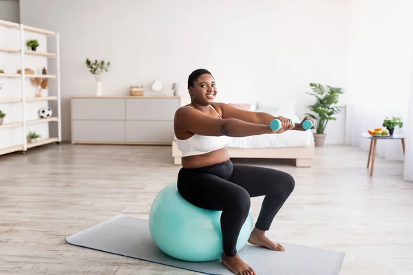 Además tamaño mujer negra haciendo ejercicio con pesas en la pelota de fitness en casa, espacio vacío —  Fotos de Stock