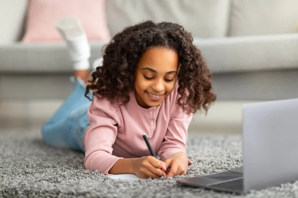 A fazer os trabalhos de casa. Menina americana africana feliz estudando on-line a partir de casa tomar notas na frente do laptop, deitado no tapete — Fotografia de Stock