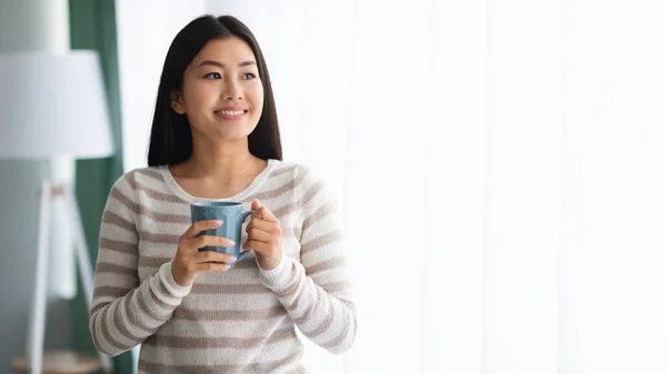 ยิ้มสาวเอเชียดื่มกาแฟในขณะที่ยืนอยู่ใกล้หน้าต่างที่บ้าน — ภาพถ่ายสต็อก