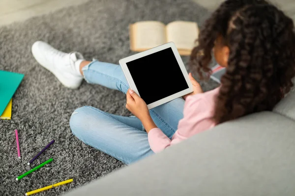 E-learning concept. African american schoolgirl using digital tablet with empty screen, sitting on floor with books