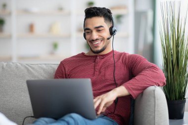 İnternetteki web sitesine katılan mutlu Arap adam, dizüstü bilgisayar ve kulaklık kullanıyor.
