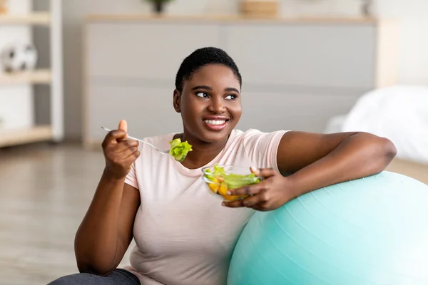 Sports and healthy nutrition for weight loss. Curvy black lady leaning on fitness ball, eating vegetable salad at home