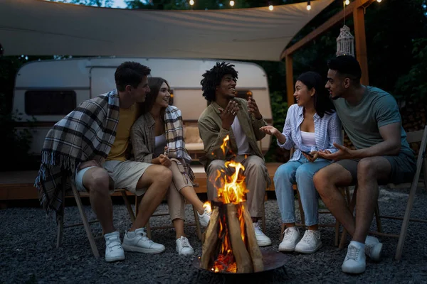 Amis multiethniques assis à côté du feu de joie, avoir une conversation amusante, passer du temps sur le voyage de camping avec RV ensemble — Photo