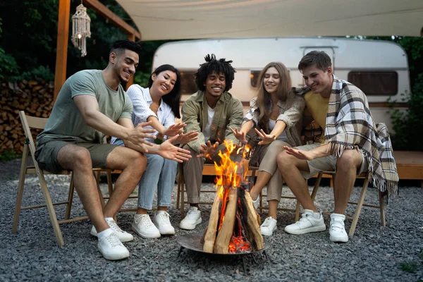 Joyeux divers jeunes amis se réchauffant les mains près du feu de camp en soirée, assis près du camping-car pendant le voyage de camping — Photo