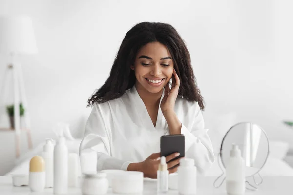 Schoonheidsapplicatie concept. glimlachende Afrikaans-Amerikaanse dame met behulp van smartphone in de slaapkamer, het dragen van witte badjas — Stockfoto