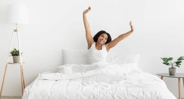Bonjour, bonne humeur et vitalité. Heureuse femme afro-américaine assise sur un lit confortable et étirant, panorama — Photo