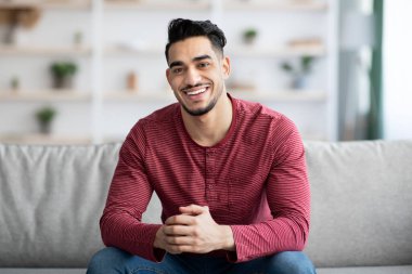 Yakışıklı Arap adam evde takılıyor, kameraya gülümsüyor.