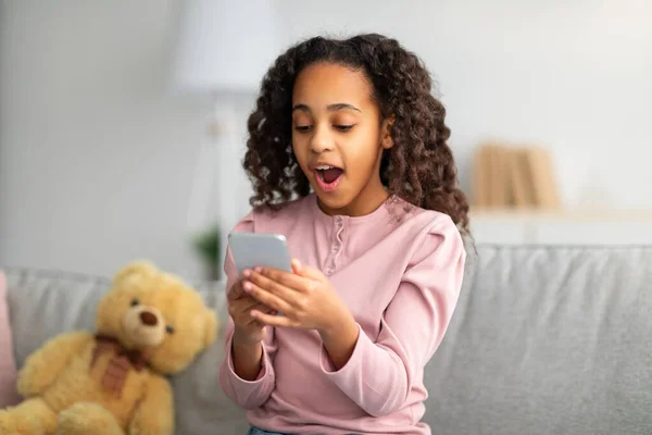 Conteúdo chocante. Surpreendida afro-americana olhando para smartphone com boca aberta, recebendo boas notícias — Fotografia de Stock