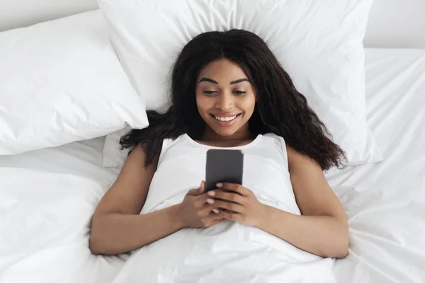 Mensaje romántico por la mañana. Feliz joven afroamericana dama acostada en la cama bajo una manta blanca y el uso de teléfono inteligente —  Fotos de Stock