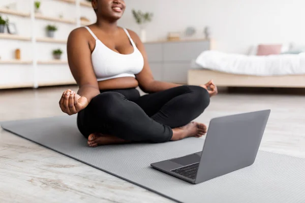 Plus size black woman having online mediatation or yoga class, sitting in lotus pose next to laptop at home, cropped view — стоковое фото