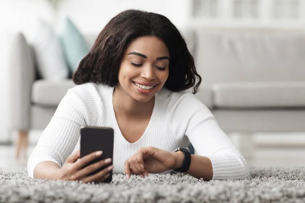 Mujer afroamericana feliz usando smartwatch con notificador de correo electrónico y teléfono inteligente, descansando en casa —  Fotos de Stock