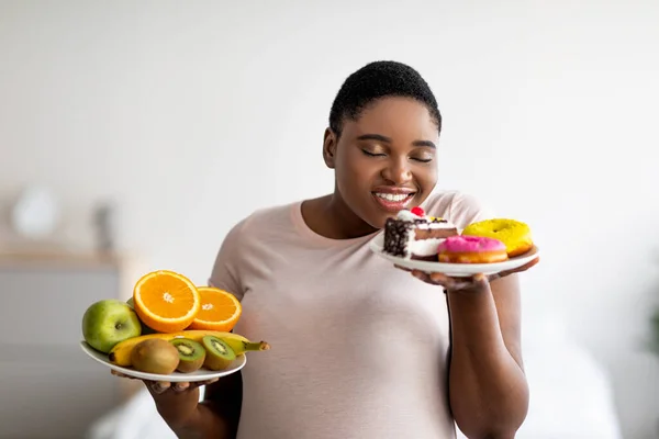 Fyllig svart kvinna som håller tallrikar med frukt och sötsaker, luktar desserter, frestad att bryta viktminskning kost hemma — Stockfoto