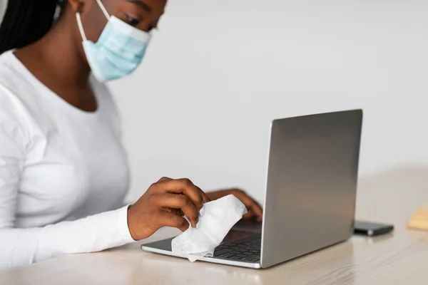 Black woman wearing medical mask cleaning laptop keyboard