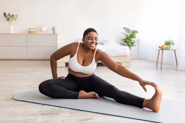 Señora negra curvilínea estirando las piernas, haciendo ejercicio en la esterilla de yoga, llevando un estilo de vida activo, tratando de perder peso en casa —  Fotos de Stock