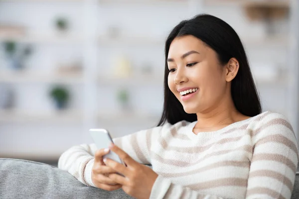 Lächelnde junge Asiatin surft im Internet auf dem Smartphone, während sie zu Hause ruht — Stockfoto