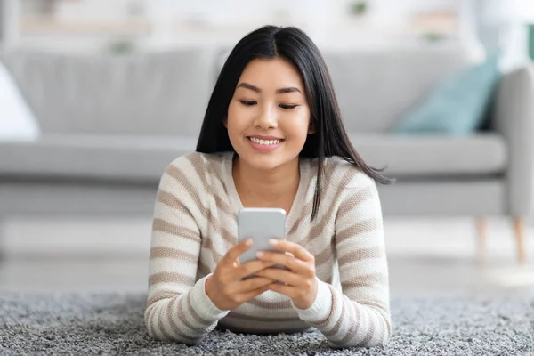 Kommunikation online. Glad asiatisk kvinna vilar på golvet med smartphone — Stockfoto