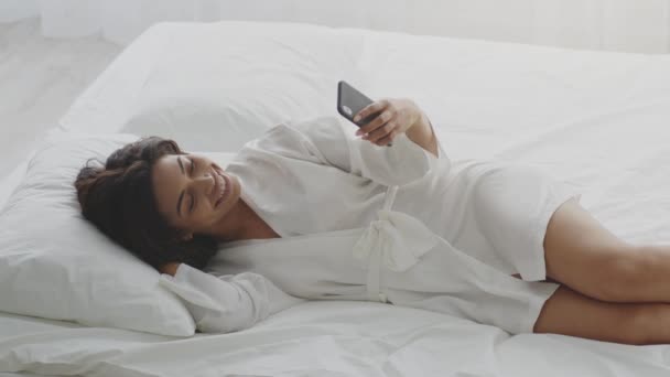 若いアフリカ系アメリカ人女性がスマートフォンで自撮りし、ソーシャルネットワークに投稿し、ベッドに横たわっています — ストック動画